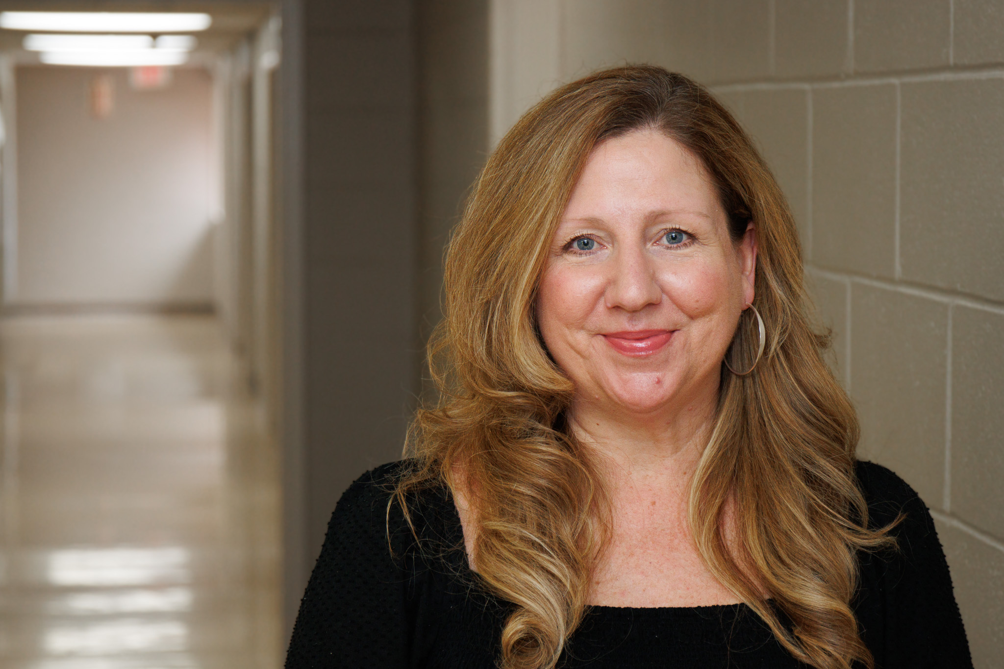 Portrait of a smiling woman with long blonde hair and blue eyes, standing in a beige hallway.
